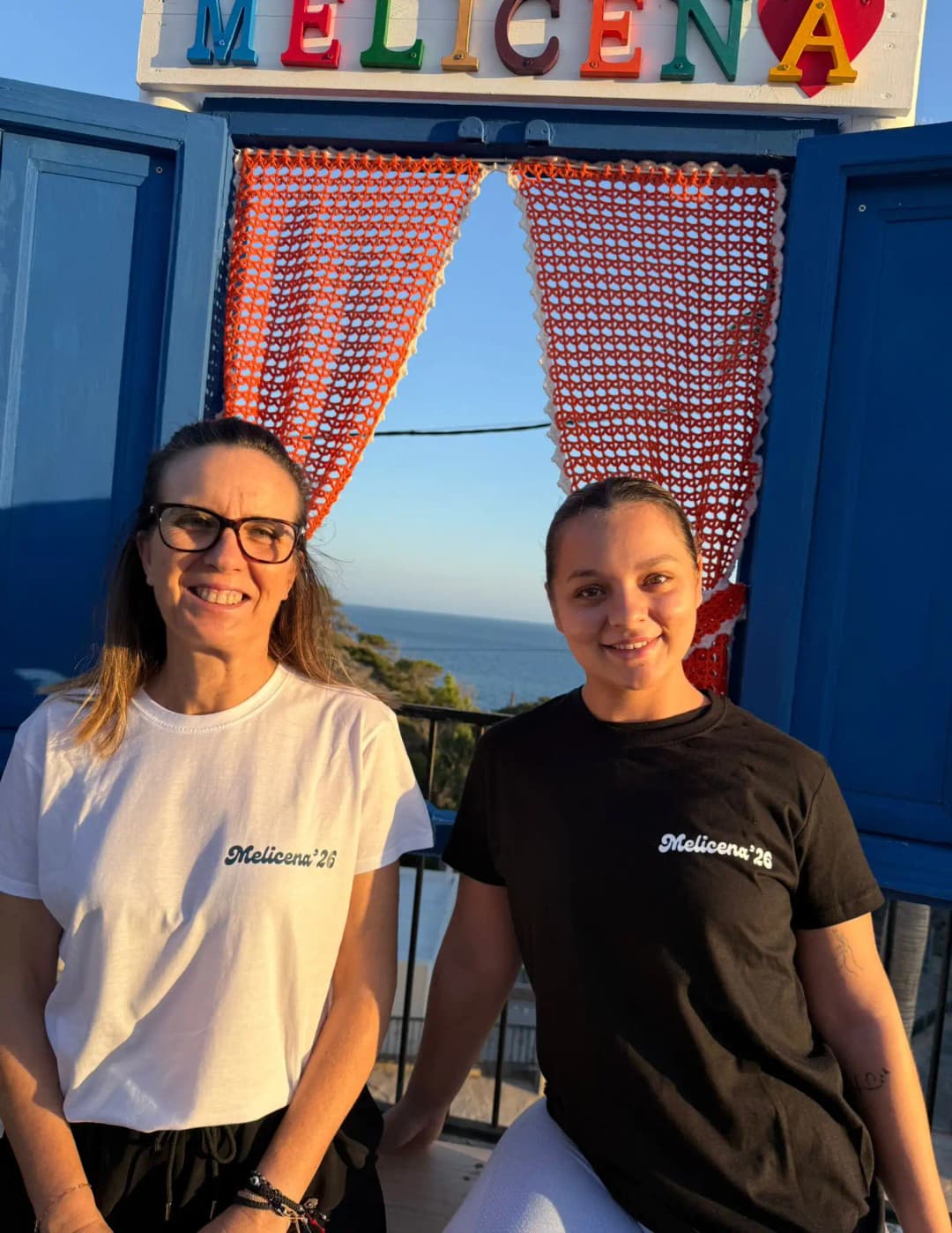 Chicas con las camisetas oficiales de Melicena 2026, frontal. Tenemos la versión en blanco y negro, ambas ponen 'Melicena 2026'.